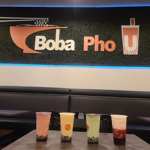 Four colorful boba drinks on a table in front of the Boba Pho U restaurant sign.