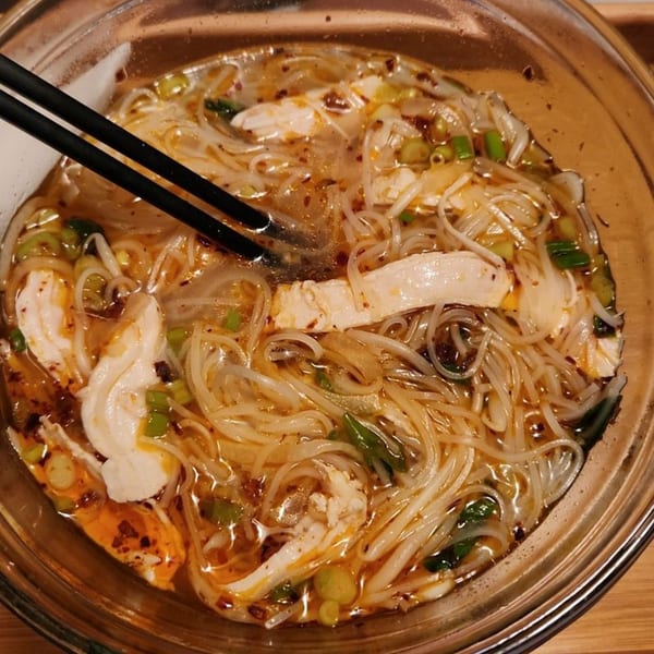 A close-up of a bowl of Vietnamese pho with chicken, noodles, and green onions, with chopsticks.